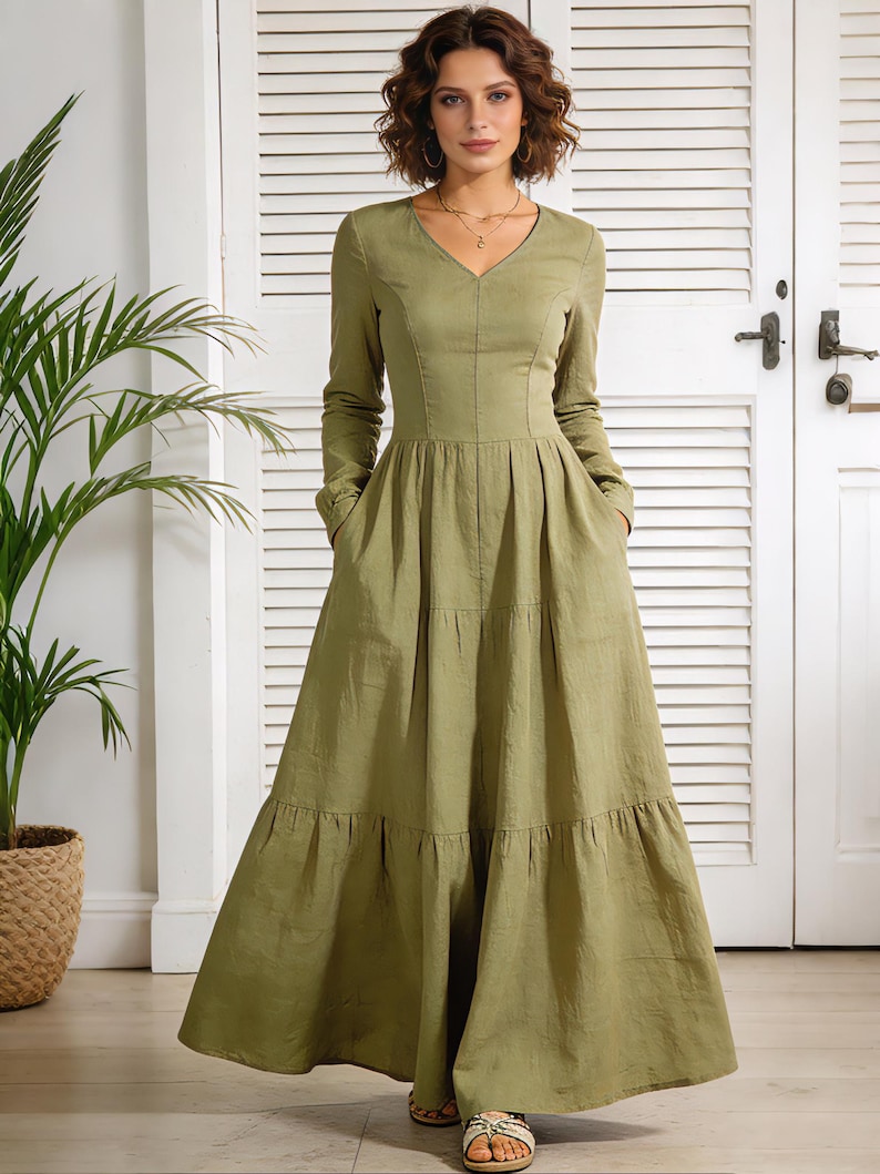 Op de afbeelding: Een olijfgroene maxi-jurk met lange mouwen en een V-hals. De jurk heeft een gelaagde rok, zakken en een getailleerd lijfje. Het model draagt sandalen. De achtergrond omvat een witte deur en een plant in een pot.