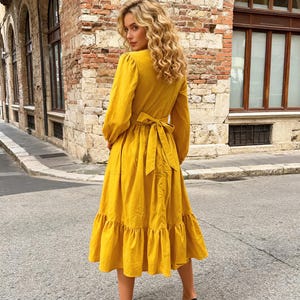 May include: A long-sleeved, yellow midi dress with a V-neck and a tie-back waist. The dress features a tiered ruffle hem and is paired with black high heels. The model is standing on a paved street with brick buildings in the background.
