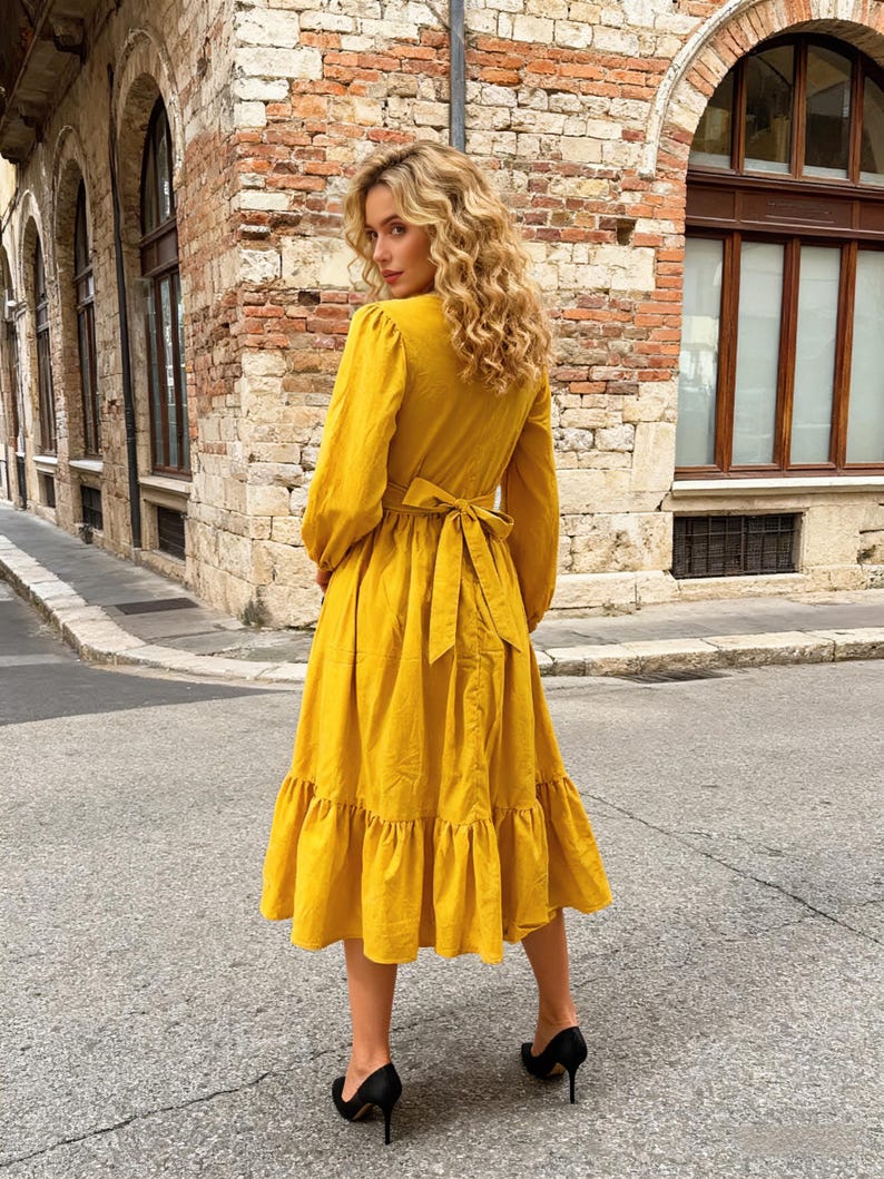 May include: A long-sleeved, yellow midi dress with a V-neck and a tie-back waist. The dress features a tiered ruffle hem and is paired with black high heels. The model is standing on a paved street with brick buildings in the background.