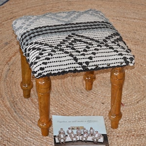 May include: A small, square stool with a patterned top in shades of grey and black, supported by four turned wooden legs. The stool sits on a woven jute rug. A card with text and a group of people is in front of the stool.