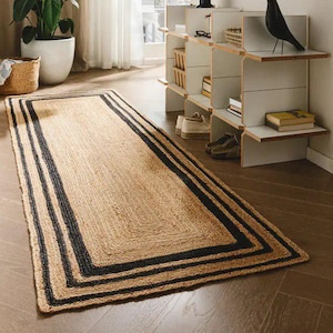 May include: A rectangular jute rug in natural tan, edged with three black stripes. The rug is on a wooden floor. The room includes a large potted plant and a decorative shelf, creating a warm, inviting atmosphere.