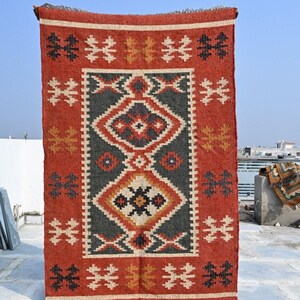 May include: A rectangular, hand-woven rug with a geometric design. The rug features a central pattern of diamonds and other shapes in shades of gray, red, and cream. The border and background are primarily red. The rug is displayed outdoors.