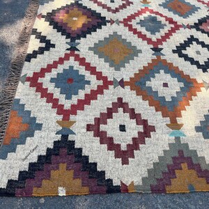 May include: A rectangular rug with a geometric diamond pattern in shades of red, blue, gold, and gray on a cream background. The rug has a fringed edge and is made of woven material.