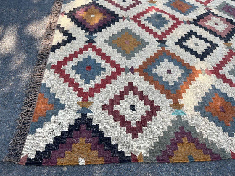 May include: A rectangular rug with a geometric diamond pattern in shades of red, blue, gold, and gray on a cream background. The rug has a fringed edge and is made of woven material.