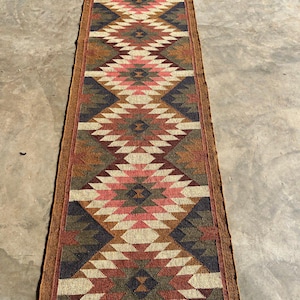 May include: A long, rectangular runner rug with a geometric diamond pattern. The rug features a color palette of red, pink, brown, beige, and navy blue. The rug is made of woven material and has a brown border.