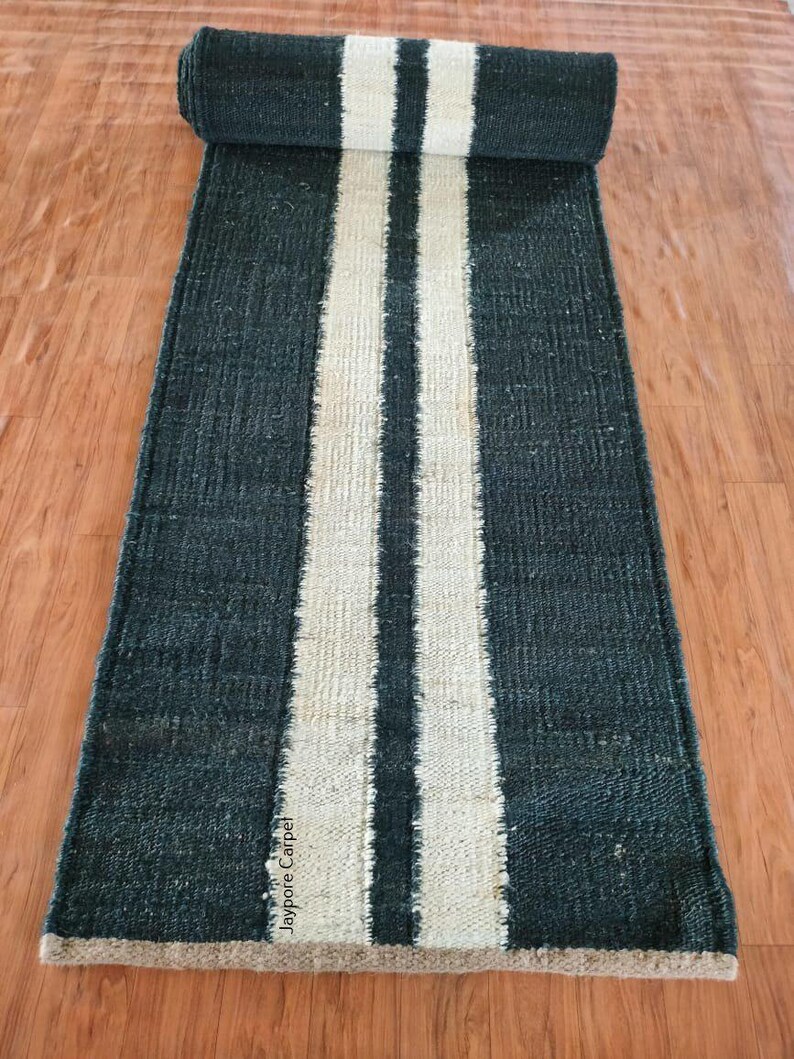 May include: A rolled-up, dark teal runner rug with two wide, cream-colored stripes down the center. The rug is made of a textured material and is displayed on a wooden floor. The rug has a rectangular shape.