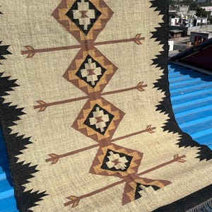 May include: A beige and black woven rug with geometric diamond and arrow designs. The rug features a repeating pattern of brown and black diamonds with arrow-like extensions. The edges are trimmed with a black zigzag pattern and fringe.