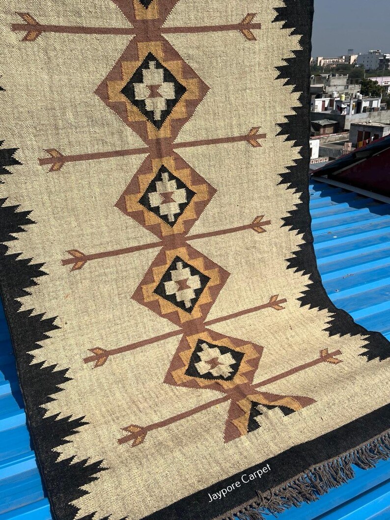 May include: A beige and black woven rug with geometric diamond and arrow designs. The rug features a repeating pattern of brown and black diamonds with arrow-like extensions. The edges are trimmed with a black zigzag pattern and fringe.