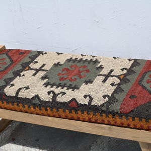 May include: Wooden bench with a rectangular seat featuring a colorful, patterned textile. The textile incorporates red, green, black, and beige geometric designs. The bench has a natural wood frame.