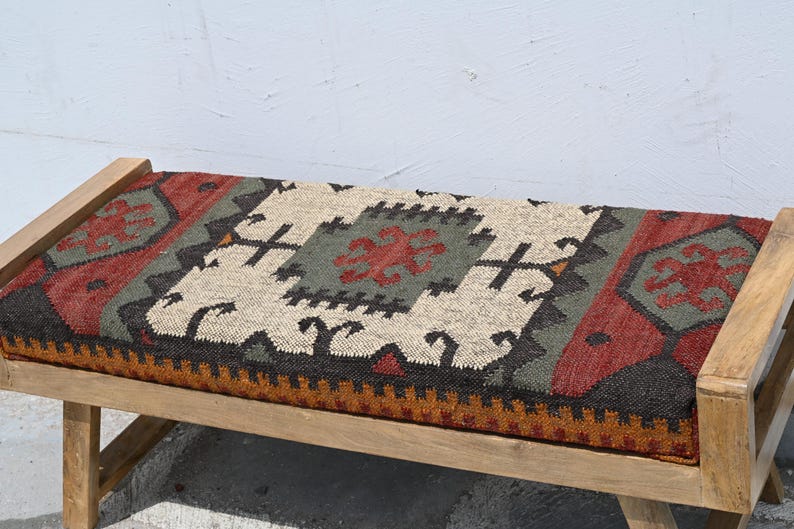May include: Wooden bench with a rectangular seat featuring a colorful, patterned textile. The textile incorporates red, green, black, and beige geometric designs. The bench has a natural wood frame.