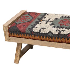 May include: A wooden bench with a rectangular seat featuring a colorful, patterned textile. The textile incorporates red, gray, black, and white geometric designs. The bench has a light-colored wooden frame and angled legs.