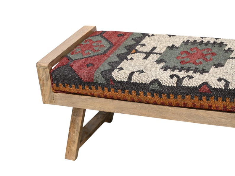 May include: A wooden bench with a rectangular seat featuring a colorful, patterned textile. The textile incorporates red, gray, black, and white geometric designs. The bench has a light-colored wooden frame and angled legs.