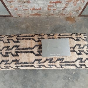 Op de afbeelding: Een rechthoekig tapijt met een beige en zwart geometrisch patroon. Een zilveren laptop ligt erop. De tekst "Jaypore Carpet" is zichtbaar op de laptop.
