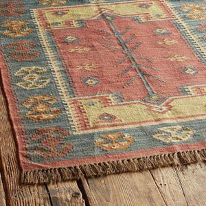 May include: A rectangular rug with a central red field featuring a stylized tree design. The rug has a blue border with repeating geometric patterns and a beige inner border. The rug has a fringed edge and is placed on a wooden surface.