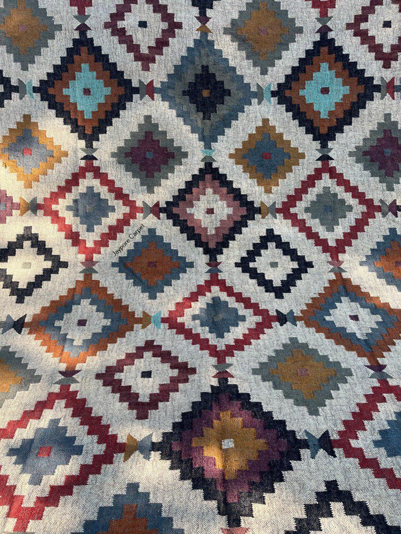 May include: A close-up of a patterned rug with a geometric design. The rug features a repeating diamond pattern in shades of red, blue, teal, brown, and gold on a neutral background. The text "Jaypore Carpet" is visible.