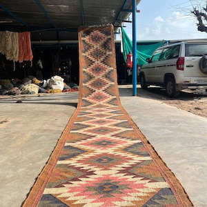 May include: A long, rectangular rug with a geometric pattern in shades of brown, pink, blue, and cream. The rug has a fringed edge and is displayed outdoors on a concrete surface. The design features diamond and zigzag shapes.