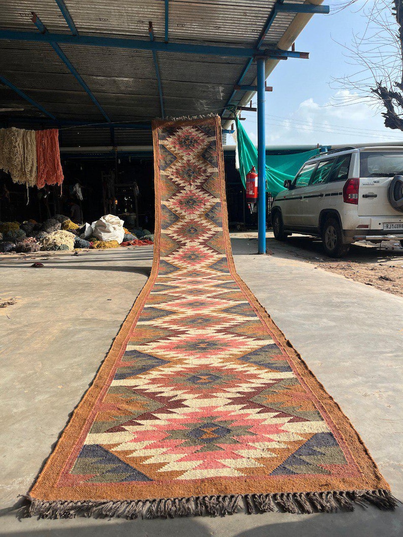 May include: A long, rectangular rug with a geometric pattern in shades of brown, pink, blue, and cream. The rug has a fringed edge and is displayed outdoors on a concrete surface. The design features diamond and zigzag shapes.
