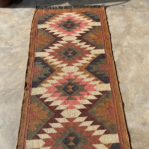 May include: A rectangular runner rug with a geometric diamond pattern. The rug features a color palette of brown, beige, pink, and navy blue. The rug has a fringed edge and is likely made of woven material.