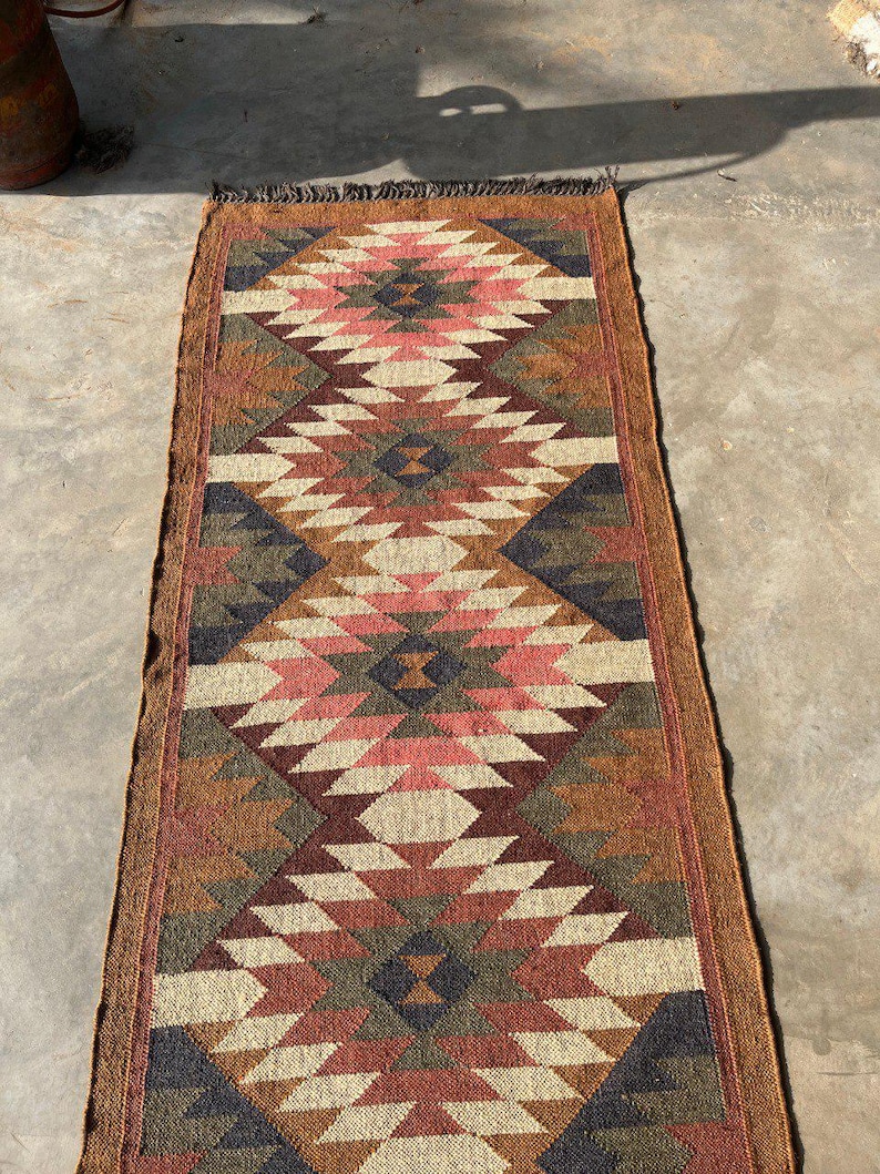 May include: A rectangular runner rug with a geometric diamond pattern. The rug features a color palette of brown, beige, pink, and navy blue. The rug has a fringed edge and is likely made of woven material.