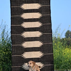 May include: A black and beige patterned rug with a geometric design is displayed outdoors. The rug has a series of horizontal beige shapes against a black background. The text "Jaypore Carpet" is visible at the top. A small dog is in front of the rug.