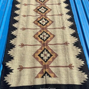 May include: Rectangular beige rug with a geometric pattern in brown, black, and gold. The rug has a black border with a zigzag design and fringe. The text "Jaypore Carpet" is visible on the rug.