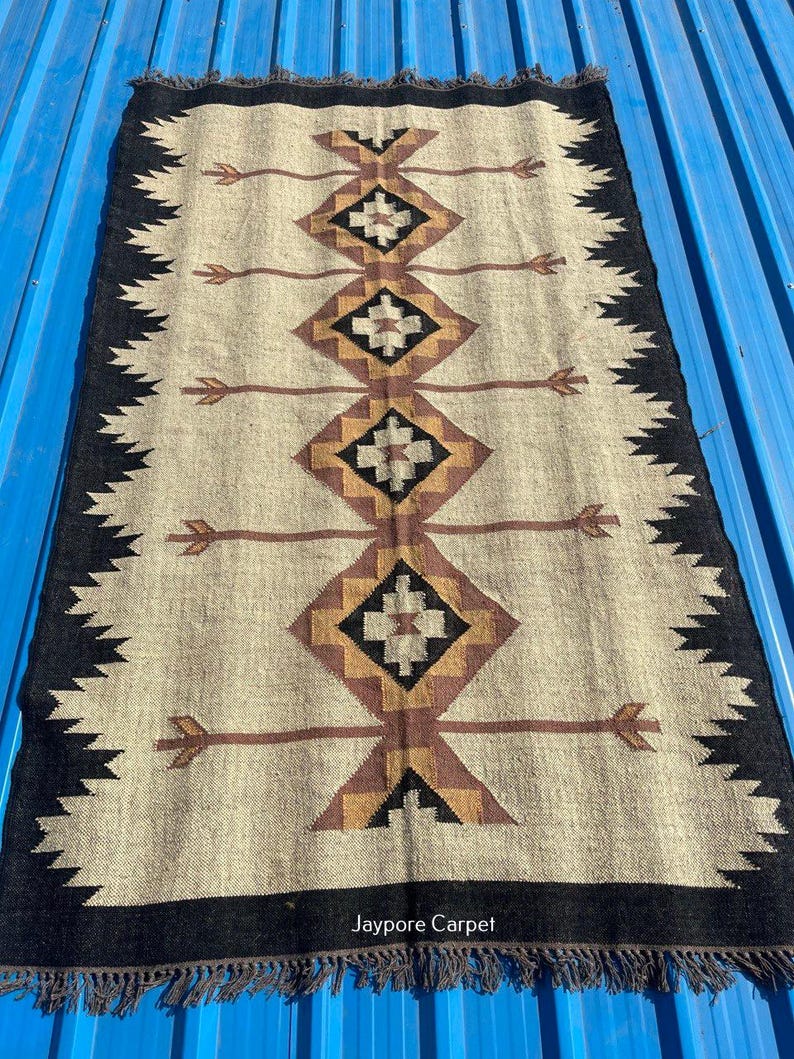 May include: Rectangular beige rug with a geometric pattern in brown, black, and gold. The rug has a black border with a zigzag design and fringe. The text "Jaypore Carpet" is visible on the rug.