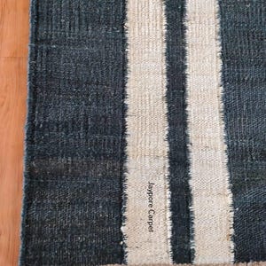 May include: A rectangular rug with a dark blue background and two vertical off-white stripes. The rug has a textured appearance and a beige border. The text "Jaypore Carpet" is visible on the rug.