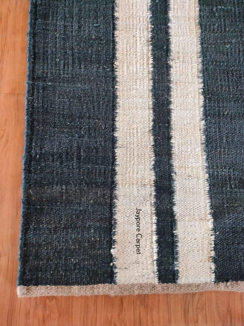 May include: A rectangular rug with a dark blue background and two vertical off-white stripes. The rug has a textured appearance and a beige border. The text "Jaypore Carpet" is visible on the rug.