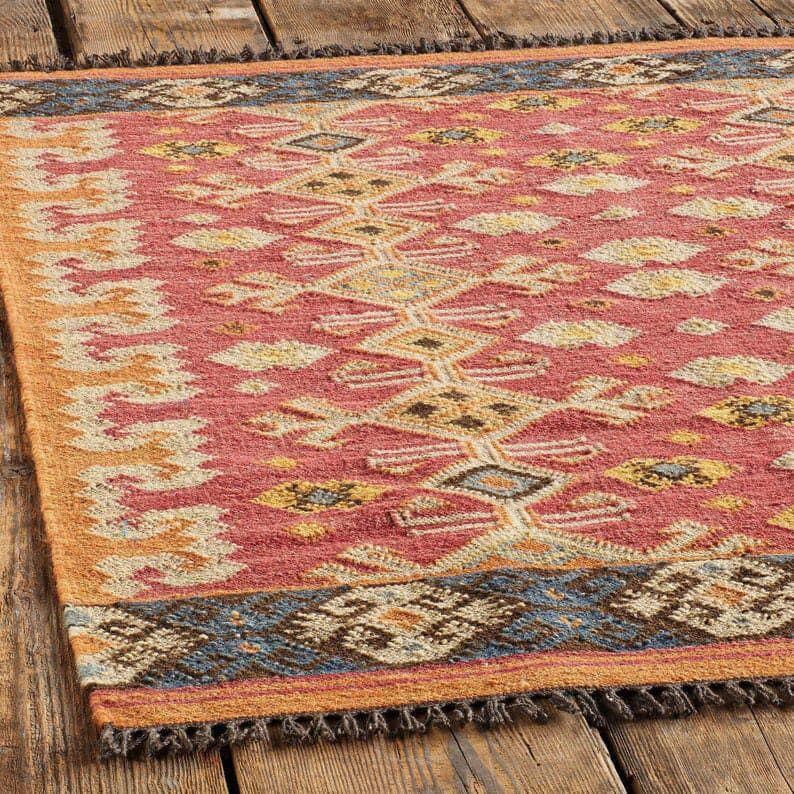 May include: A rectangular rug with a geometric pattern in shades of red, yellow, blue, and brown. The rug has a fringed edge and is placed on a wooden surface. The design features diamond and zigzag shapes.