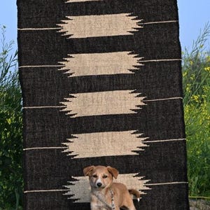 May include: A black and beige patterned rug with the text "Jaypore Carpet" is displayed outdoors. A small brown dog stands in front of the rug, attached to a chain. The rug has a geometric design.