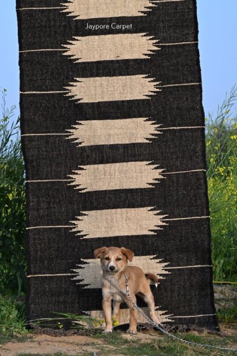 May include: A black and beige patterned rug with the text "Jaypore Carpet" is displayed outdoors. A small brown dog stands in front of the rug, attached to a chain. The rug has a geometric design.