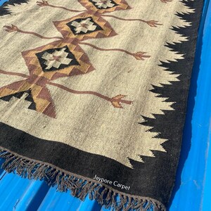 May include: A rectangular beige rug with a geometric pattern in brown, black, and gold. The rug has a black border with a fringe and the text "Jaypore Carpet".