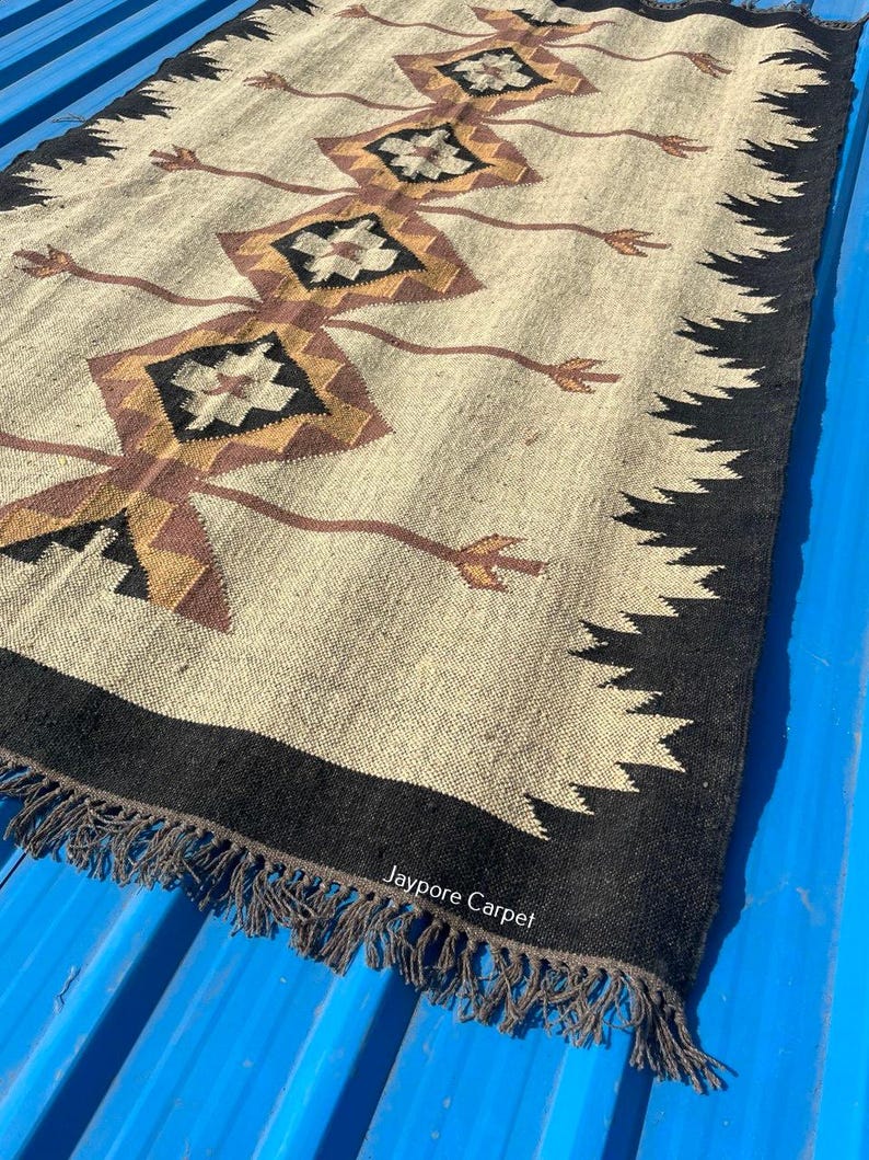 May include: A rectangular beige rug with a geometric pattern in brown, black, and gold. The rug has a black border with a fringe and the text "Jaypore Carpet".