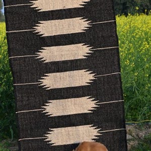 May include: A black and beige rectangular rug with a geometric design. The rug features horizontal beige shapes with zigzag edges on a black backdrop. The text "Jaypore Carpet" is visible. A dog is near the rug.
