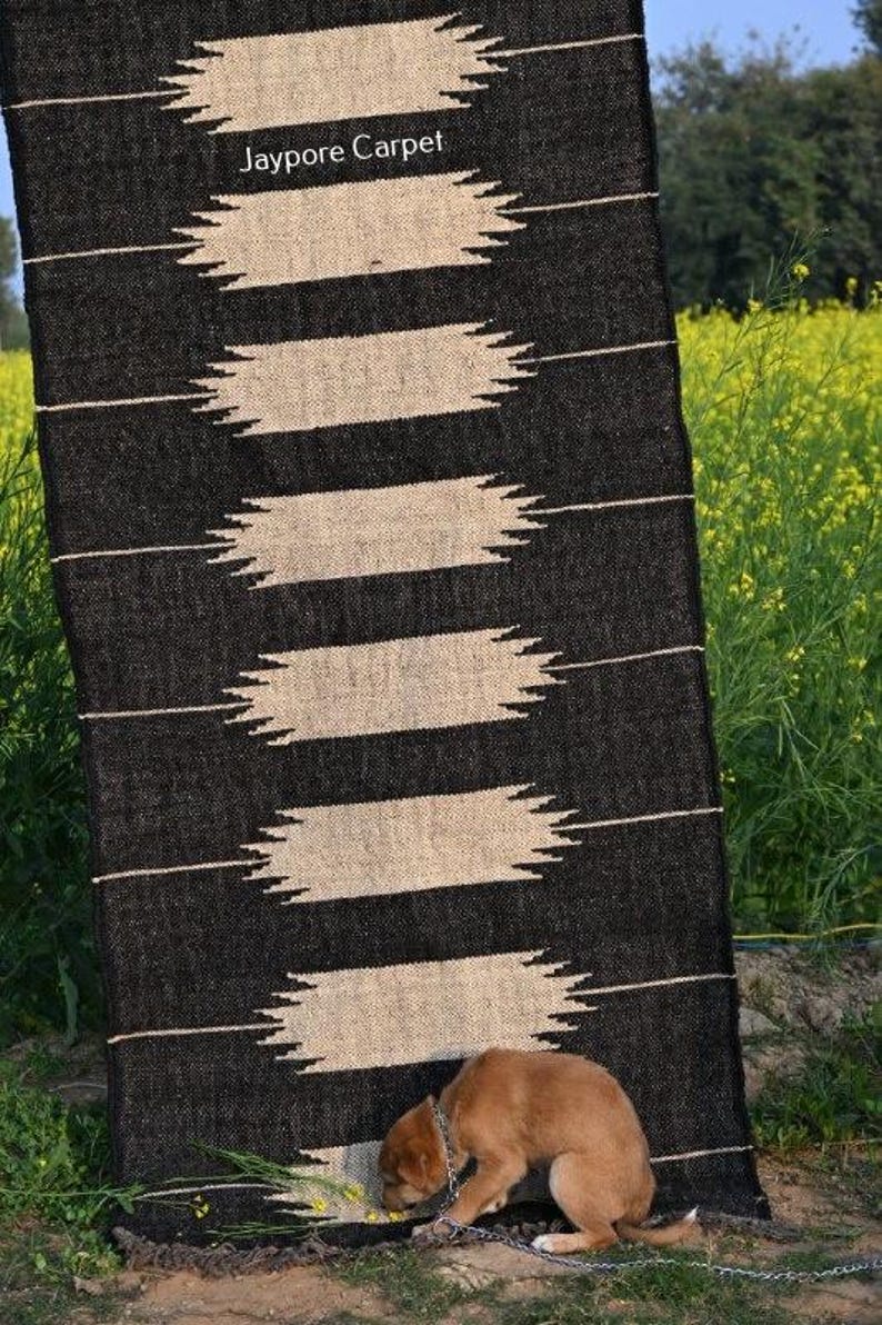 May include: A black and beige rectangular rug with a geometric design. The rug features horizontal beige shapes with zigzag edges on a black backdrop. The text "Jaypore Carpet" is visible. A dog is near the rug.