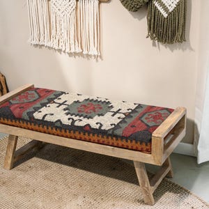 May include: A wooden bench with a rectangular seat featuring a patterned textile in red, black, beige, and green. The bench has a light wood frame and angled legs, suitable for home decor.