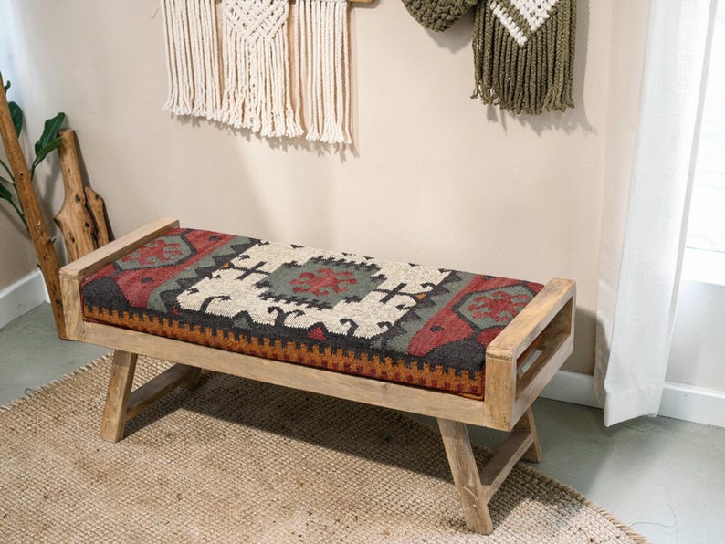 May include: A wooden bench with a rectangular seat featuring a patterned textile in red, black, beige, and green. The bench has a light wood frame and angled legs, suitable for home decor.