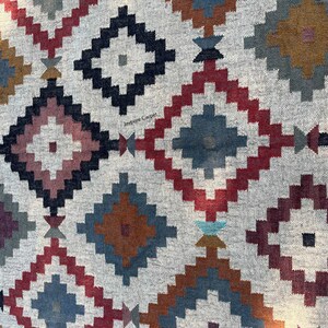 May include: A close-up of a gray rug with a geometric diamond pattern. The diamonds are in shades of red, blue, brown, and black. The rug has a textured appearance and is likely made of woven fabric.