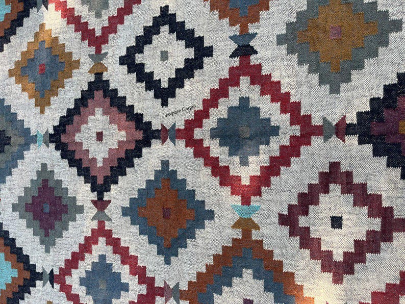 May include: A close-up of a gray rug with a geometric diamond pattern. The diamonds are in shades of red, blue, brown, and black. The rug has a textured appearance and is likely made of woven fabric.