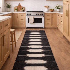 May include: A black and white patterned runner rug with fringe, in a kitchen with wooden cabinets and a white stove. The rug features a geometric design and the text "Jaypore Carpet".