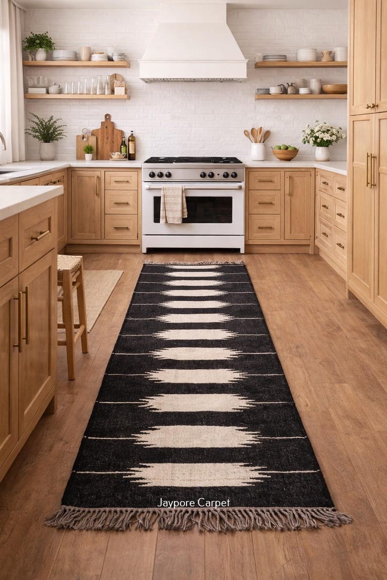 May include: A black and white patterned runner rug with fringe, in a kitchen with wooden cabinets and a white stove. The rug features a geometric design and the text "Jaypore Carpet".