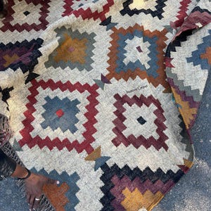 May include: A woven rug with a geometric pattern in shades of gray, red, blue, and gold. The rug features a repeating square design with a fringed edge. The text "jaypore Carpet" is visible on the rug.