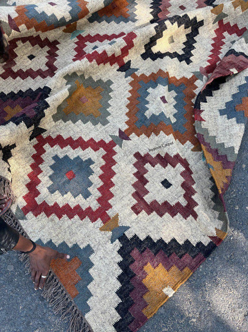 May include: A woven rug with a geometric pattern in shades of gray, red, blue, and gold. The rug features a repeating square design with a fringed edge. The text "jaypore Carpet" is visible on the rug.