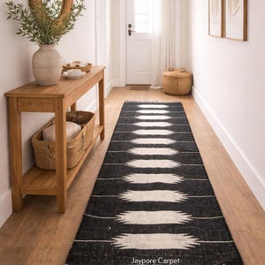May include: A black and white patterned runner rug with a fringed edge. The rug has a repeating geometric design. The text "Jaypore Carpet" is visible. The rug is in a hallway with a wooden console table and a mirror.