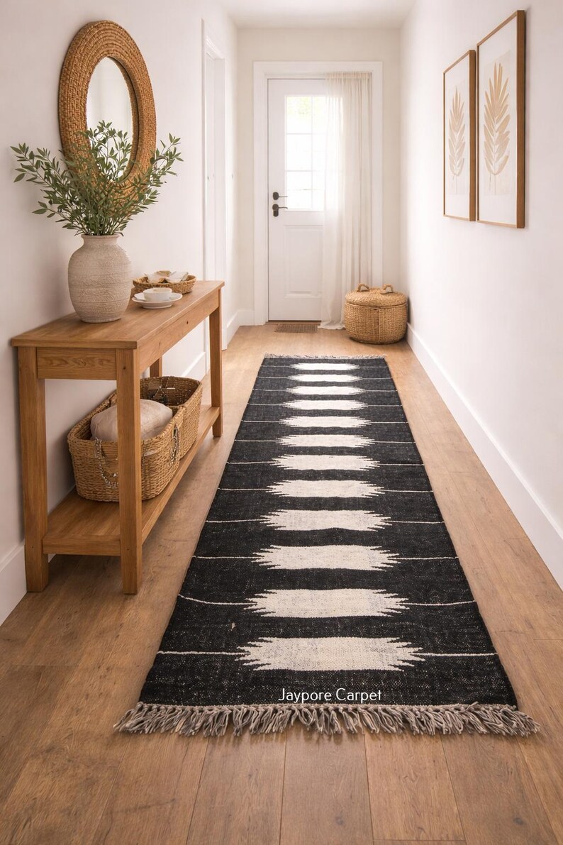 May include: A black and white patterned runner rug with a fringed edge. The rug has a repeating geometric design. The text "Jaypore Carpet" is visible. The rug is in a hallway with a wooden console table and a mirror.