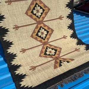 May include: Beige and brown geometric patterned rug with a black border and fringe. The rug features diamond and arrow designs in shades of brown and black. The rug is made of a woven material.