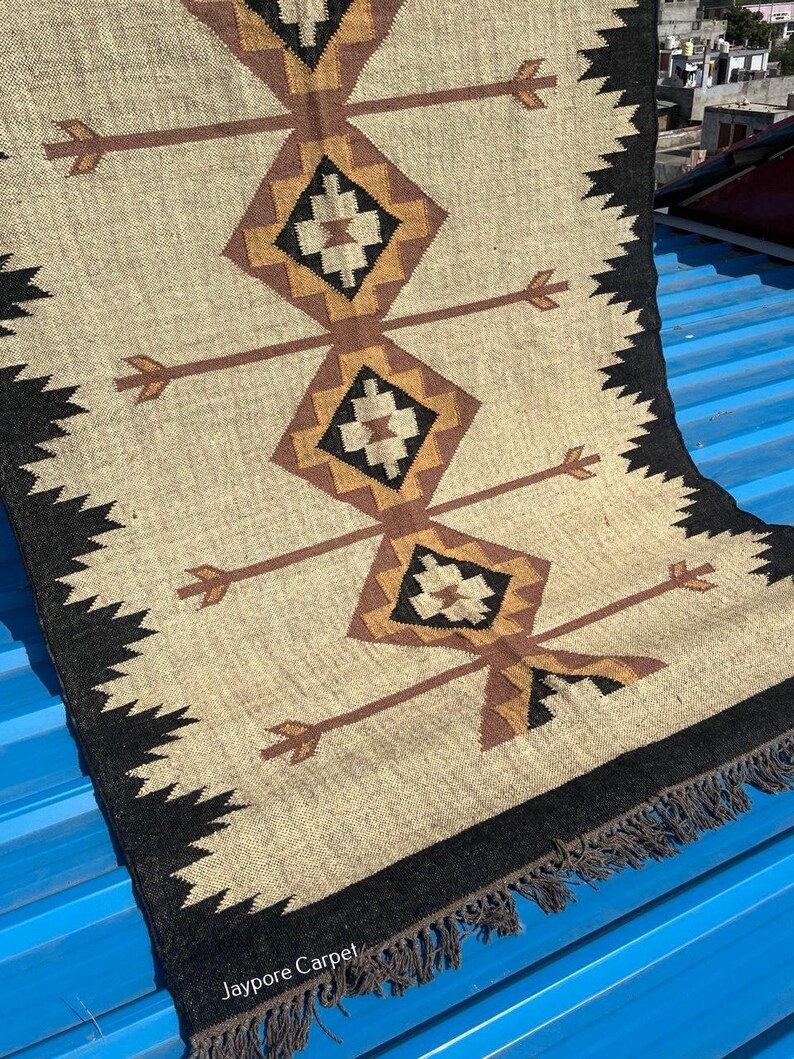 May include: Beige and brown geometric patterned rug with a black border and fringe. The rug features diamond and arrow designs in shades of brown and black. The rug is made of a woven material.