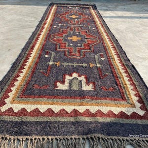 May include: A rectangular, dark blue rug with a geometric pattern in red, yellow, and white. The rug has a fringed edge and the text "Jaypore Carpet" is visible in the bottom right corner. The rug is likely made of woven material.