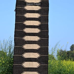 May include: A black and beige woven rug with a geometric pattern. The rug features horizontal beige shapes on a black backdrop. The text "Jaypore Carpet" is visible. The rug is hanging outdoors against a blue sky.