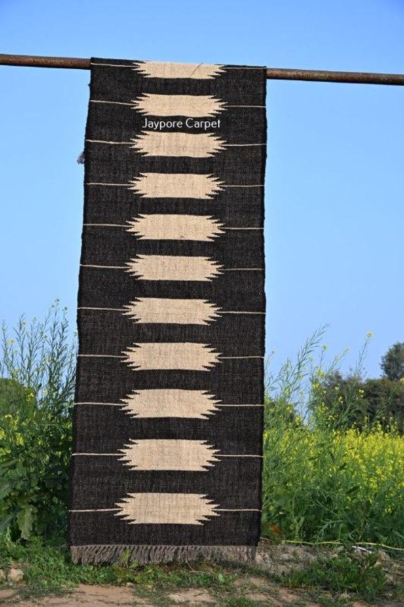 May include: A black and beige woven rug with a geometric pattern. The rug features horizontal beige shapes on a black backdrop. The text "Jaypore Carpet" is visible. The rug is hanging outdoors against a blue sky.
